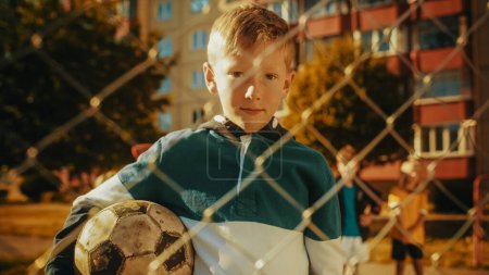 Portrait of a Cute Boy in Green and White Hoodie Holding a Soccer Ball on an Outdoors Field in the Neighborhood. Young Football Player Looking at