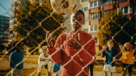 Téléchargez les photos : Portrait d'un jeune beau garçon noir lançant et attrapant un ballon de football tout en regardant la caméra et souriant. Sporty African Kid rêve sur - en image libre de droit