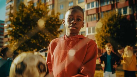 Téléchargez les photos : Portrait d'un jeune beau garçon noir tenant un ballon de football tout en regardant la caméra et en souriant. African Kid debout dans la cour avec - en image libre de droit
