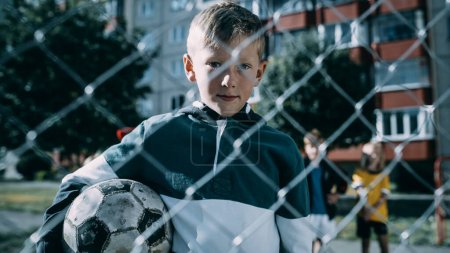 Téléchargez les photos : Portrait d'un garçon mignon tenant un ballon de soccer sur un terrain extérieur dans le quartier. Jeune footballeur regardant la caméra, souriant. Images - en image libre de droit