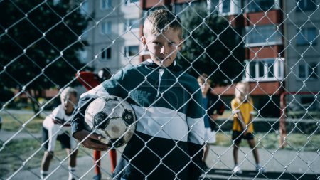 Téléchargez les photos : Portrait d'un garçon mignon tenant un ballon de soccer sur un terrain extérieur dans le quartier. Jeune footballeur regardant la caméra, souriant. Images - en image libre de droit