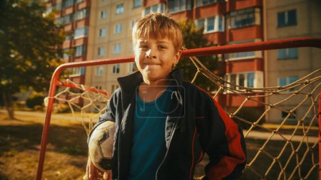 Téléchargez les photos : Portrait d'un beau garçon portant un manteau de sport tenant un ballon de football dans le quartier. Jeune footballeur regardant la caméra, souriant. Debout - en image libre de droit