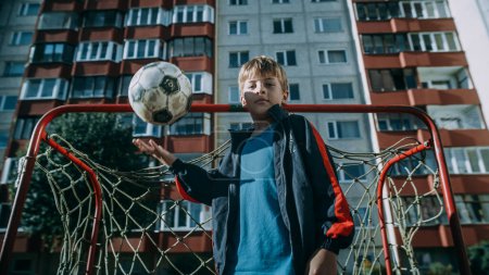 Téléchargez les photos : Portrait d'un jeune garçon portant des vêtements de sport lançant et attrapant un vieux ballon de soccer sur un terrain extérieur dans une cour arrière. Jeune footballeur regardant - en image libre de droit