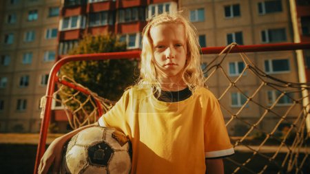 Téléchargez les photos : Portrait d'une fille blonde sérieuse en T-shirt jaune tenant un ballon de football dans le quartier, debout à côté d'une porte d'objectif. Jeune joueur de football - en image libre de droit