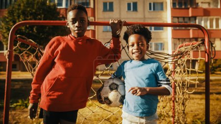 Téléchargez les photos : Portrait de deux jeunes beaux garçons noirs debout ensemble tout en regardant la caméra et souriant. Frère plus jeune tenant un ballon de football. Enfants africains - en image libre de droit