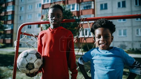 Téléchargez les photos : Portrait de deux jeunes garçons africains talentueux debout ensemble tout en regardant caméra et sourire. Frère aîné tenant un ballon de football. Froid - en image libre de droit