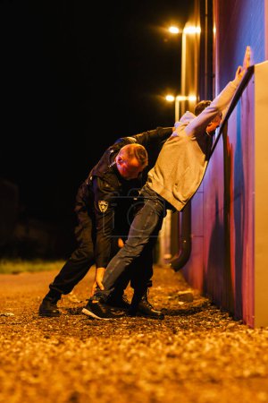 A Professional Middle Aged Policeman Performing a Pat-Down Search on a Fellon in Empty Back Alley. Documentary-like Shot of Proper Procdure of