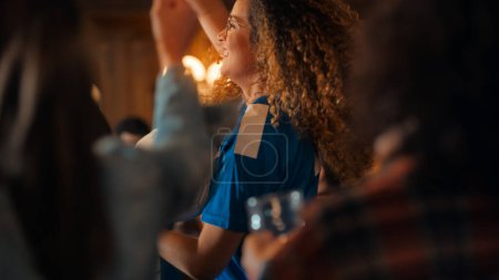 Porträt einer multiethnischen jungen Frau im blauen Trikot, die in einer Menge von Sportfans in einem Pub jubelt und Fernsehübertragungen verfolgt. Freunde feiern
