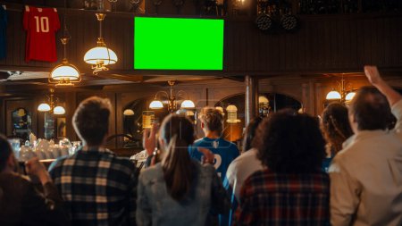 Gruppe multikultureller Freunde, die ein Live-Sportspiel im Fernsehen mit Green-Screen-Bildschirm in einer Bar verfolgen. Glückliche Fans jubeln und schreien, feiern