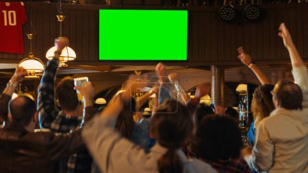 Gruppe multikultureller Freunde, die ein Live-Sportspiel im Fernsehen mit Green-Screen-Bildschirm in einer Bar verfolgen. Glückliche Fans jubeln und schreien, feiern