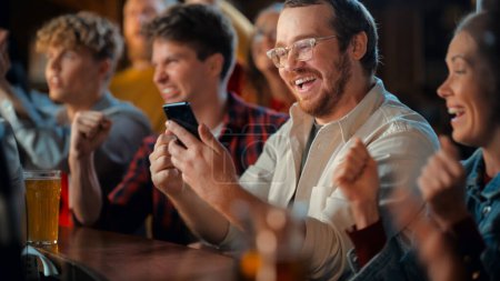 Excited Man in Glasses Sitting at a Bar Stand, Using a Smartphone, Feeling Alive After Putting a Bet on His Favorite Soccer Team. Ecstatic When