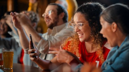 Excited Female Holding a Smartphone, Sitting at a Bar Stand. Nervous About the Sports Bet She Put on a Her Favorite Soccer Team. Joyful When Football