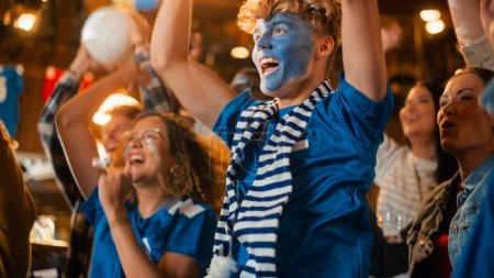 Nahaufnahme Porträt eines hübschen jungen Fußballfans mit gemaltem blau-weißem Gesicht, der in einer Bar in einer Menschenmenge steht, singt, springt und jubelt.