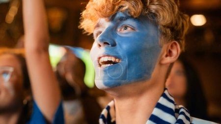 Close Up Portrait of a Handsome Young Soccer Fan with Painted Blue and White Face Standing in a Crowd in a Bar, Chanting, Jumping, Cheering for a