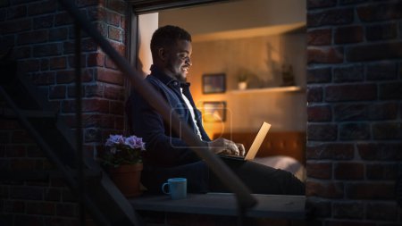 Schöner, lächelnder, schwarzafrikanischer Mann arbeitet am Laptop, während er zu Hause auf der Fensterbank sitzt. Porträt eines jungen männlichen Freiberuflers, der an einem Computer arbeitet