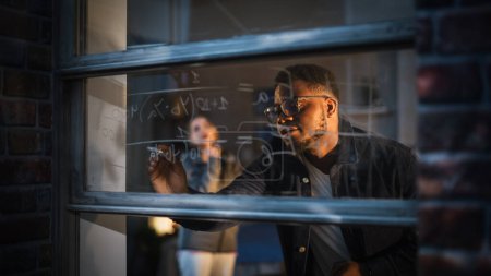 Schwarzer junger Mann erklärt einer Freundin eine mathematische Gleichung an einem Fenster zu Hause, indem er löschbare Glasmarker verwendet. Zwei multiethnische Universitäten
