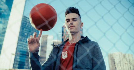 Porträt eines hübschen jungen Fußballspielers, der in die Kamera blickt und einen roten Fußballball an seinem Finger dreht. Stilvoller Fußballer steht hinter einem Zaun