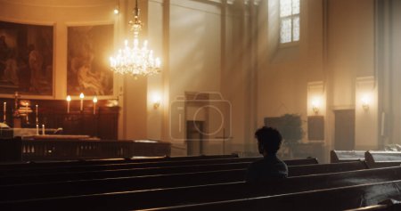 Téléchargez les photos : Le jeune chrétien est assis pieusement dans une église majestueuse, les mains pliées, il cherche conseil auprès de la foi et de la spiritualité tout en priant. Croyance religieuse - en image libre de droit