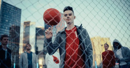 Porträt eines hübschen jungen Fußballspielers, der in die Kamera blickt und einen roten Fußballball an seinem Finger dreht. Stilvoller Fußballer steht hinter einem Zaun