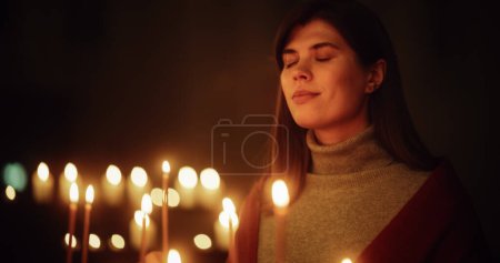 Cinematic Portrait of a Young Woman Standing Next to Lit Candles with Closed Eyes and a Smile. Female Christian Finding Inner Peace in Religion