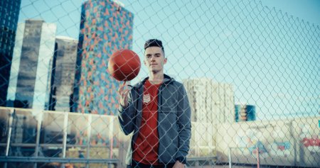 Porträt eines stilvollen jungen Fußballspielers, der für die Kamera posiert und einen roten Ball am Finger dreht. Schöner Fußballer, der in einer Stadt hinter einem Zaun steht