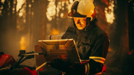 Porträt eines Feuerwehrmannes in Sicherheitsausrüstung mit Hochleistungs-Laptop-Computer, der über eine Situation mit einem gefährlichen Waldbrand berichtet. Schwerer Tag