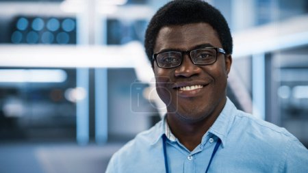 Téléchargez les photos : Beau Africain portant des lunettes et une chemise bleue souriant et regardant la caméra. Jeune homme noir travaillant comme ingénieur ou scientifique en technologie - en image libre de droit
