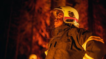 Mutiger junger afroamerikanischer Feuerwehrmann in Sicherheitsuniform und Helm, der sich bei einem Waldbrand im Wald umsieht. Profi-Kader