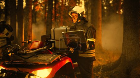 Mutiger, geschickter Feuerwehrmann, der neben einem ATV steht und Laptop-Computer im Wald mit wütendem Reisigfeuer benutzt. Superintendent oder Einsatzleiter sorgen für Sicherheit