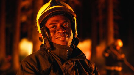 Nahaufnahme des Porträts eines tapferen jungen erwachsenen Feuerwehrmannes in Sicherheitsuniform und Helm, der während eines Flächenbrandes für die Kamera posiert. Professionelle