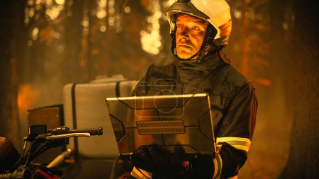Portrait of a Handsome Fireman in Safety Gear Using Heavy-Duty Laptop Computer, Reporting on a Situation with a Dangerous Wildland Fire in a Forest