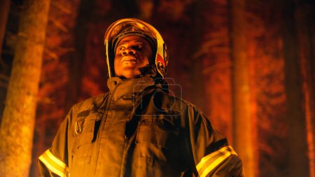 Mutiger junger afroamerikanischer Feuerwehrmann in Sicherheitsuniform und Helm, der sich bei einem Waldbrand im Wald umsieht. Profi-Kader