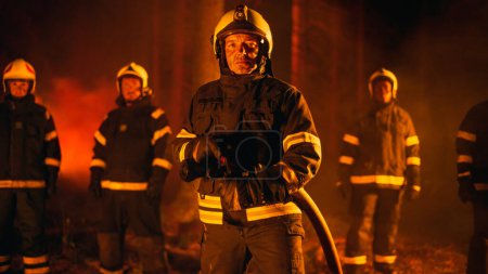 Porträt eines tapferen, gut aussehenden Mannschaftsführers in Sicherheitsuniform und Helm, der während eines Flächenbrandes mit Feuerwehrleuten für die Kamera posiert. Berufsfeuerwehrmann