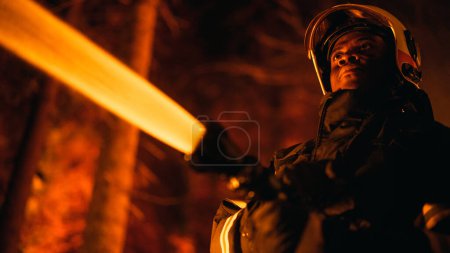 Close Up Low Angle Creative Portrait eines tapferen afroamerikanischen Feuerwehrmannes, der einen Waldbrand mit einem Feuerwehrschlauch bekämpft. Schwarzer Feuerwehrmann hält hoch