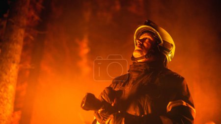 Erfahrener Feuerwehrmann löscht einen Waldbrand tief im Wald. Profi in Schutzuniform und Helm mit Feuerwehrschlauch, der nach oben schaut