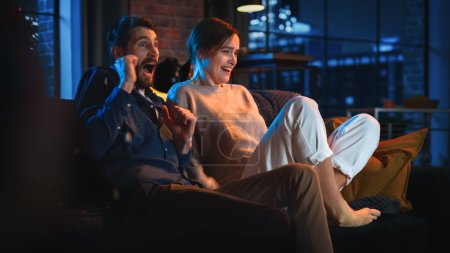 Portrait of Beautiful Couple Spending Time at Home, Sitting on a Couch, Watching Scary TV Show in Their Stylish Loft Apartment. Man and Woman