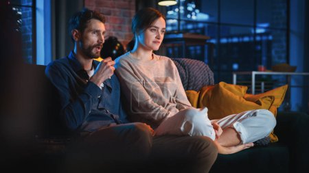 Portrait of Beautiful Couple Spending Time at Home, Sitting on a Couch, Watching Scary TV Show in Their Stylish Loft Apartment. Man and Woman