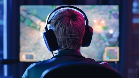 Parte posterior del ángulo principal de una joven jugadora que usa auriculares y juega un videojuego en una computadora personal en una sala de estar Neon Lit en casa. Acogedor