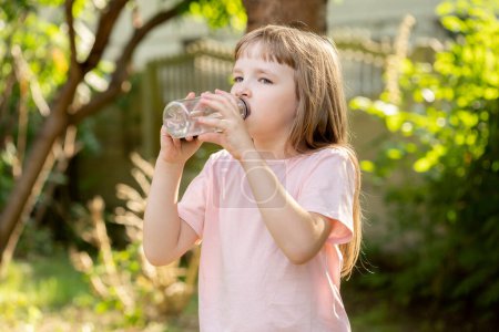 Téléchargez les photos : Elementary school age girl, child hydrating, drinking water from a transparent reusable plastic bottle outdoors, outside, one person, real people lifestyle. Sporty kid drinks water, blurry background - en image libre de droit