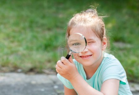 Photo for Child, young happy curious girl holding a magnifying glass next to her eye smiling, outdoors poprtrait, copy space. Smart kid with a loupe, real curiosity, science and education, discovery concept - Royalty Free Image