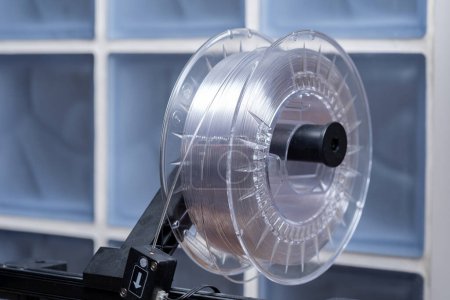 Un rollo de filamento de impresión 3D transparente blanco de alta calidad montado en un dispositivo de impresora 3D moderno, primer plano. Materiales termoplásticos de grado consumidor utilizados para la creación de prototipos y el diseño de productos objeto