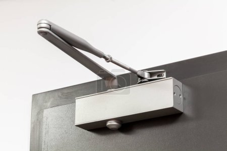 Close-up showing a modern silver door closer mechanism, with its hydraulic arm extended, installed on the upper edge of a grey house apartment building interior door in a bright setting, indoors shot