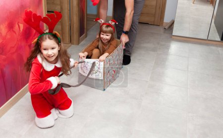 Christmas at home, two cheerful children, one dressed as Santa and the other with reindeer antlers, are playfully pulling a makeshift sleigh through the hallway of a home, simulating a festive ride