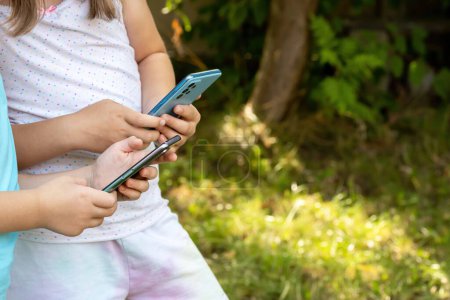 Photo for Young girls, children focusing on their smartphones, mobile phones, texting with friends or browsing the internet, using apps while standing outdoors on a bright day, copy space. Kids and technology - Royalty Free Image