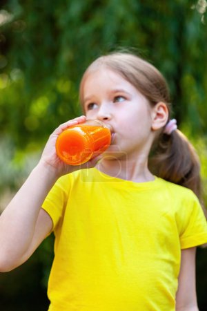 Photo for Young girl in a vibrant yellow t-shirt outdoors enjoying a refreshing drink from a clear bottle filled with orange carrot juice. Natural daylight, casual relaxed real lifestyle shot, vertical comp - Royalty Free Image