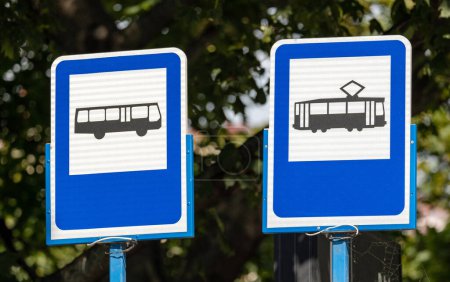 Blaue Bus- und Straßenbahnhaltestellenschilder nebeneinander, Objektaufnahme, vor verschwommenem Baumhintergrund, Tageslicht, niemand. Nahverkehr, Öko-Verkehrsmittel, abstraktes Konzept, zwei Schilder zusammen