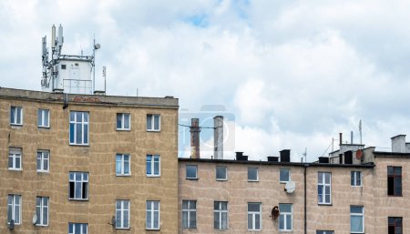 Old multi-story residential building stands under a cloudy sky. A cell tower is installed on the roof, factory power plant chimneys behind. Antennas and satellite dishes, lots of windows, apartments