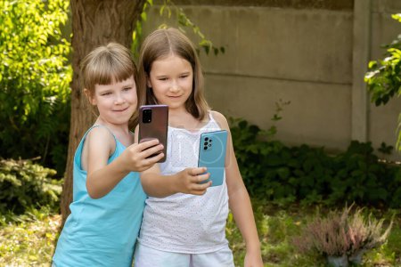 Zwei junge Mädchen, Kinder, die lächeln, während sie tagsüber ein Selfie mit ihren Smartphones im Garten machen.