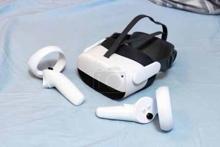 Standalone Virtual-Reality-Headset und Controller auf einem Bett zu Hause, blaue Stoffoberfläche, generische VR-Brille voller Ausstattung, Spiele und Unterhaltungserlebnisse, Cyberspace-Konzept, niemand