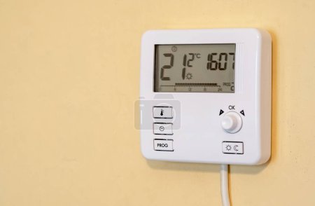Modern smart home programmable digital thermostat unit mounted on a yellow wall inside a house, showing the current room temperature and set temperature in a residential apartment setting, nobody